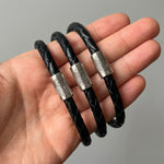 Black braided leather bracelet with silver fingerprint accent caps and a matching silver magnetic clasp, shown close-up with a finger holding it.